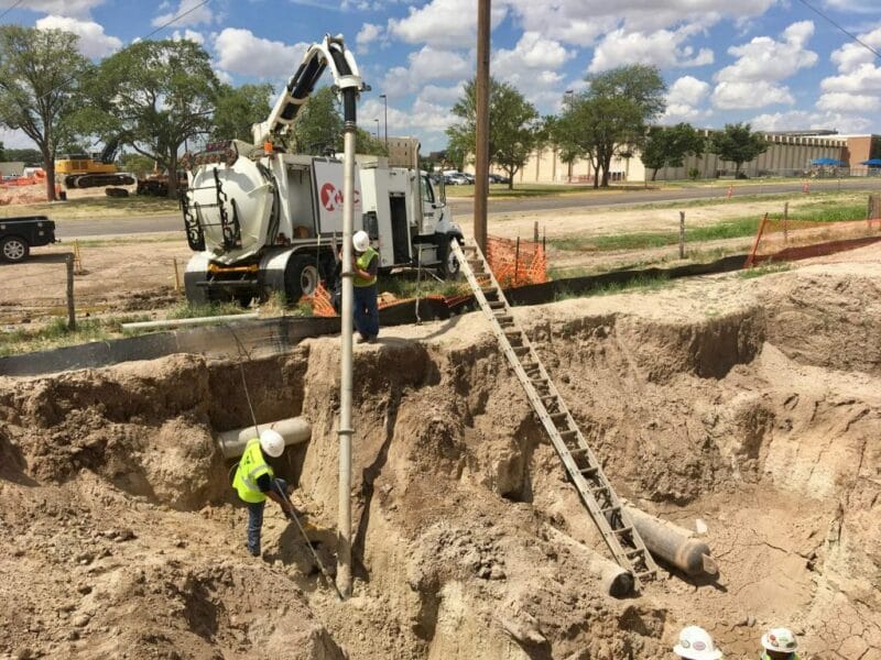 Two workers in a deep sandy excavation using an X‑Vac hydroexcavation vacuum hose and ladder beside the X‑Vac truck.