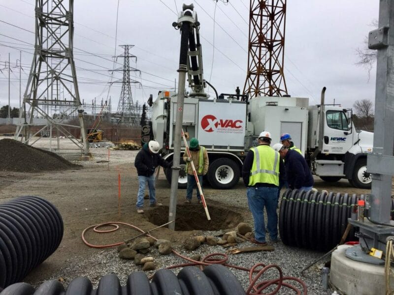Workers in high-vis vests using X-Vac hydro excavation truck and vacuum boom to dig a utility hole at a power substation.