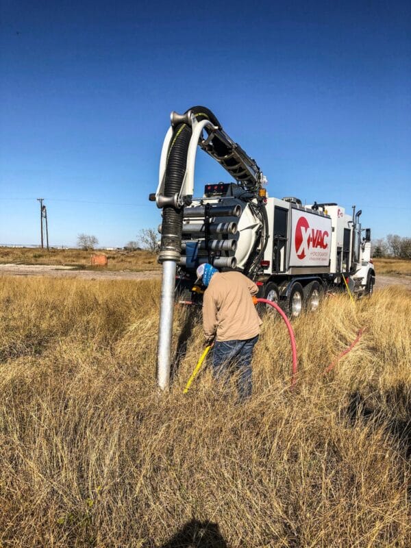 Worker handling hoses beside an X‑VAC hydroexcavation truck with boom and vacuum hose in a dry field